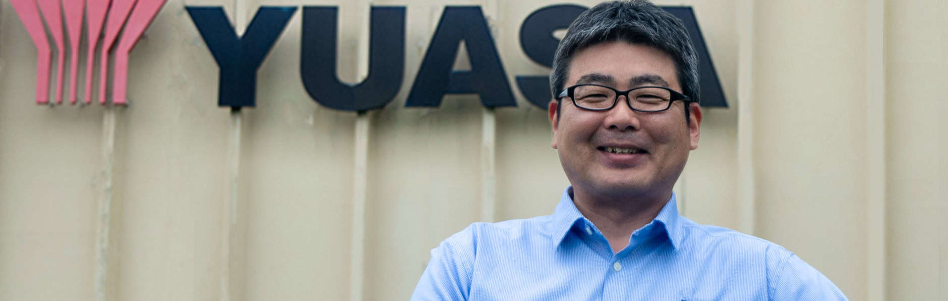 Rio Okubo, the new President and CEO of Yuasa Battery pictured in front of the Yuasa sign and logo in a blue shirt.